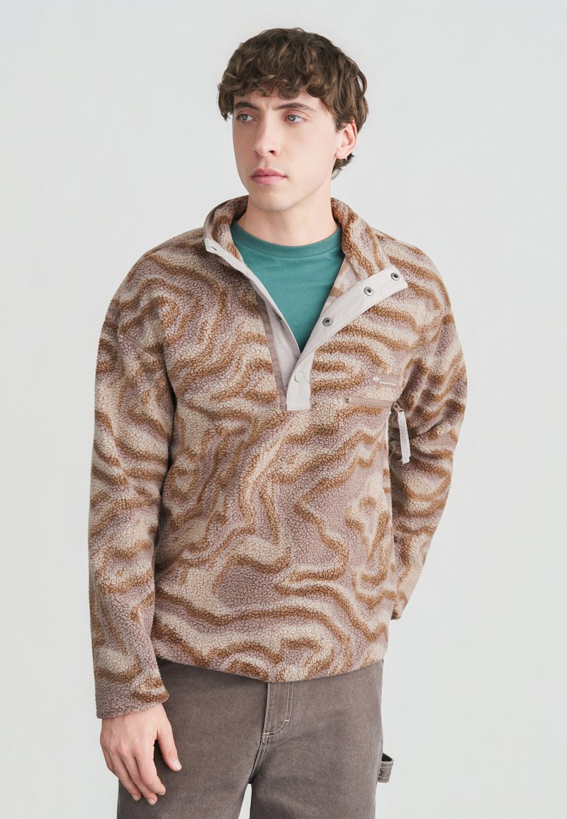 Νέος άνδρας με καφέ-ταμπά φλις μπλούζα με σχέδια πάνω σε πράσινο πουκάμισο, στέκεται με το ένα χέρι στην τσέπη μπροστά σε απλό φόντο.