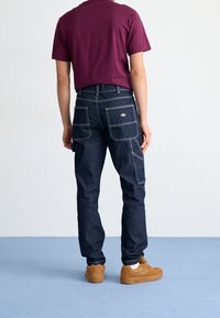 Pantalones cargo de mezclilla azul oscuro con costuras blancas contrastantes, múltiples bolsillos y un diseño ajustado, combinados con una camisa de manga corta color burdeos.