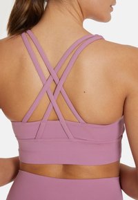 Ljubičasti sportski grudnjak s križnim naramenicama, izrađen od glatkog materijala. Ima pojas na dnu i minimalistički dizajn.