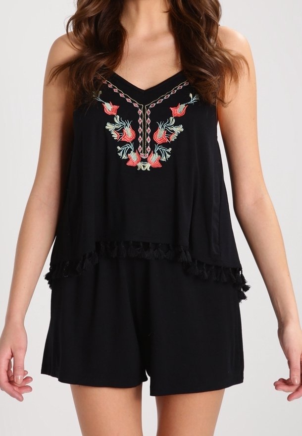 Femme portant une combinaison noire sans manches avec une broderie florale sur la poitrine et des détails de pompons le long de l'ourlet superposé.