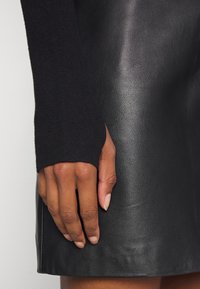 Gonna nera in pelle con una consistenza liscia, abbinata a una manica a maglia nera a coste con foro per il pollice. Posizionamento dettagliato delle mani sulla gonna.