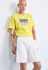 Žuti pamučni majica s grafičkim printom, uparen s bijelim kratkim hlačama s crnim bočnim prugama i Adidas logom.