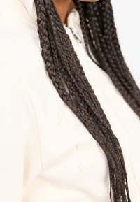 Felpa bianca con tessuto testurizzato, che presenta un design impresso in modo sottile sul davanti. Lunghe trecce cadono accanto al capo.