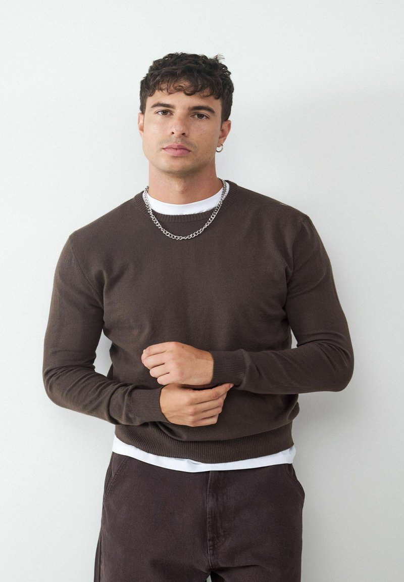 Brauner Strickpullover mit Rundhalsausschnitt, gerippten Bündchen und Saum, kombiniert mit einem weißen Unterhemd. Das Modell steht vor einem schlichten Hintergrund.