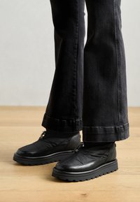 Pantofi negri slip-on cu un superior texturat, talpă din cauciuc masivă și șireturi elastice. Asortați cu blugi negri evazați care se află pe podea.