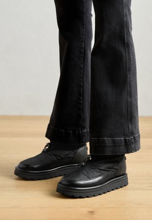 Crne slip-on cipele s teksturiranim gornjištem, masivnim gumiranim potplatom i elastičnim vezicama. Uparene s tamnim flared trapericama koje leže na podu.