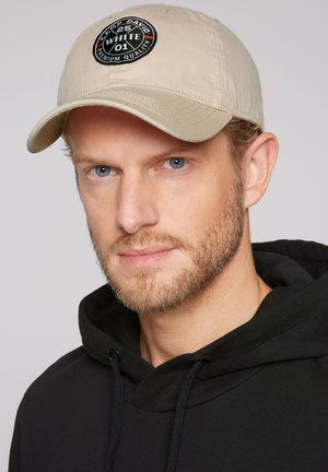 Beige cap with a round black patch featuring text and logos. Worn with a black hooded sweatshirt, emphasizing a casual style.
