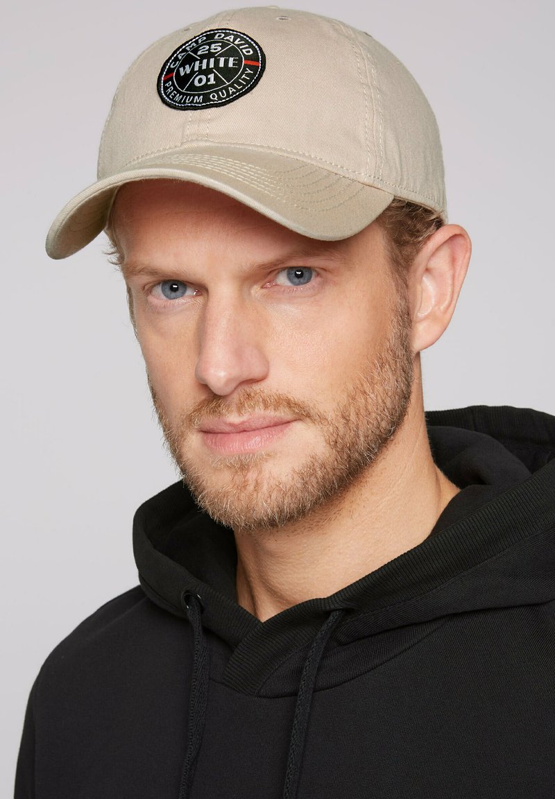 Beige cap with a round black patch featuring text and logos. Worn with a black hooded sweatshirt, emphasizing a casual style.