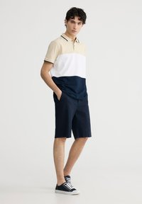 Polo a maniche corte in beige, bianco e blu navy, con strisce a contrasto e colletto. Abbinato a pantaloni corti textured blu navy fino al ginocchio e sneakers scure.