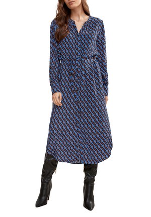 Langärmliges Kleid mit einem blauen und braunen geometrischen Muster, das eine Knopfleiste und einen Taillengurt hat, kombiniert mit schwarzen kniehohen Stiefeln.
