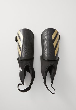 Black soccer shin guards with gold diagonal stripes, textured surface, and elastic sleeves for secure attachment.