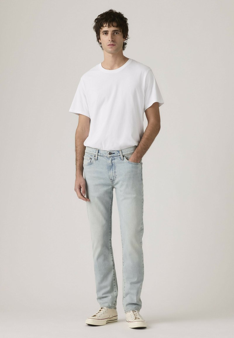 Young man standing wearing a plain white t-shirt, light blue jeans, and white sneakers against a plain background.