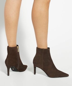 Bottines en daim marron à bout pointu avec fermetures éclair latérales et talons fins, portées sur jambes nues sur un fond clair uni.