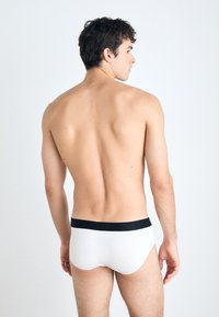 Weiße Baumwollunterhosen mit einem schwarzen Bund, von hinten an einem männlichen Modell gezeigt. Sanfte Textur, enger Sitz und einfaches Design.
