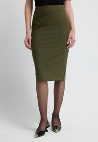 Olivenfarbene Bleistiftröcke mit glatter Textur und hoher Taille. Kombiniert mit transparenter schwarzer Strumpfhose und spitzen schwarzen Schuhen mit goldenen Akzenten.