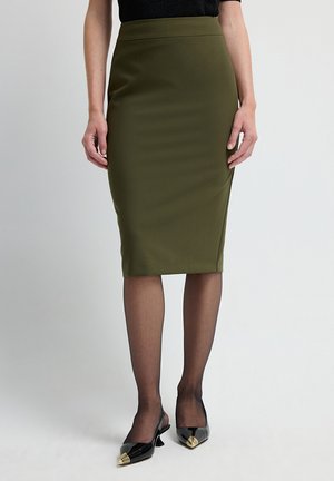 Falda lápiz verde oliva con textura suave y diseño de cintura alta. Combinada con medias negras transparentes y zapatos negros de punta con acentos dorados.