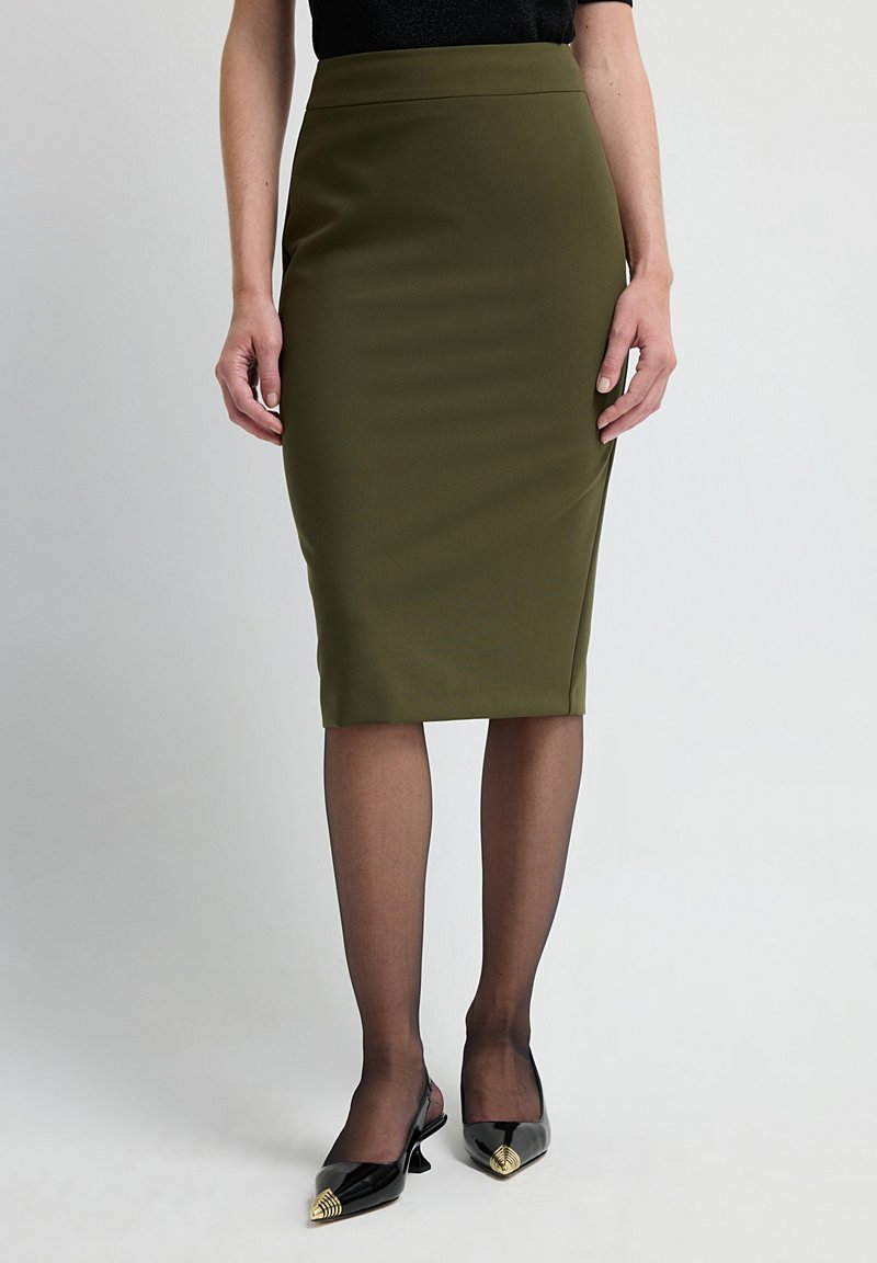 Olivenfarbene Bleistiftröcke mit glatter Textur und hoher Taille. Kombiniert mit transparenter schwarzer Strumpfhose und spitzen schwarzen Schuhen mit goldenen Akzenten.