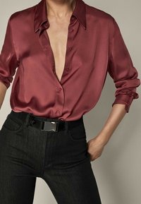 Satin bordeauxbluse med dyb V-hals og knapdetaljer, parret med højtaljede sorte jeans og et sort bælte med en blank spænde.