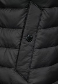 Svart vadderad jacka med en slät och mjuk textur. Har en diagonal söm och en rund svart knapp med präglad text.