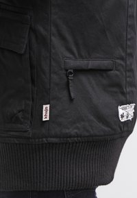 Veste noire en matière texturée, dotée d'un ourlet côtelé, de poches latérales, d'une poche zippée et d'une petite étiquette avec le nom de la marque "khujo".