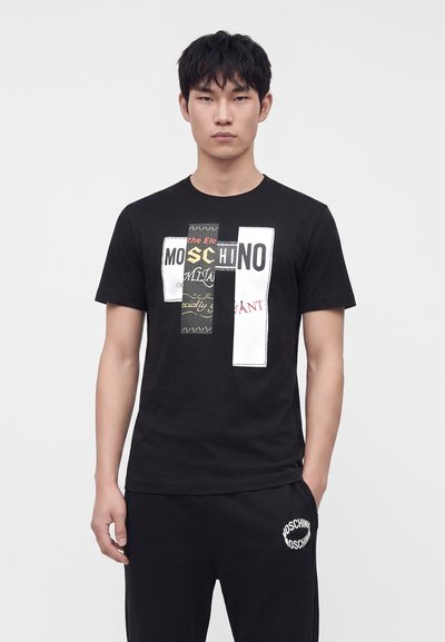 Νεαρός άνδρας φοράει μαύρο t-shirt Moschino με λογότυπο patchwork και μαύρο παντελόνι με λευκή επιγραφή Moschino κοντά στην τσέπη.