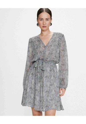 Femme portant une robe à manches longues grise à motif cachemire, avec un décolleté en V et une taille nouée, debout devant un fond blanc uni.