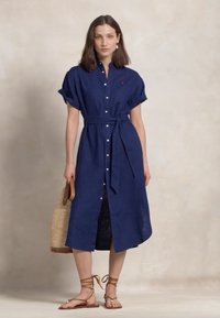 Jūras spēku zila lina kleita ar īsām uzrullētām piedurknēm, pogu priekšpusi, jostas auklu un sānu izgriezumiem; apvienota ar plakanām brūnām sandālēm un austotu somiņu.