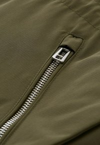 Tessuto verde oliva con una superficie leggermente texturizzata, dotato di una cerniera metallica sul lato e un design elegante e moderno.