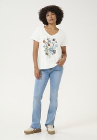 T-shirt blanc à col en V orné d'un imprimé floral coloré, associé à un jean flare bleu clair et des baskets beiges avec des accents rouges et bleus.