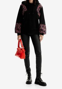 Hoodie noir avec accents rose léopard, pantalon en cuir noir et bottes noires à lacets ; tenant un sac à main rouge vif.