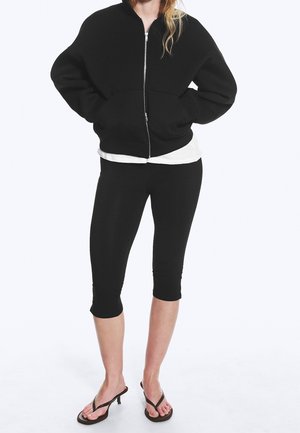Femme portant une veste noire zippée, des leggings noirs courts, une chemise blanche en dessous, et des tongs noires à talons, debout devant un fond blanc.