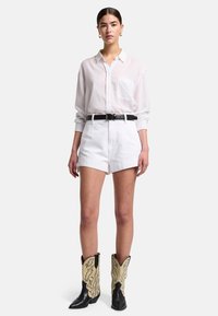 Camicia bianca a maniche lunghe con tasca, abbinata a un pantalone corto bianco e stivali da cowboy neri con motivi a forma di ala beige. Dettaglio in vita con cintura.