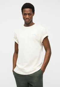 Άνδρας που φοράει λευκή μπλούζα με ρολά μανίκια και το κείμενο «Les Deux» στο στήθος, με τα χέρια στις τσέπες, στέκεται μπροστά σε απλό γκρι φόντο.