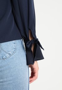 Námořnická blůza s dlouhými rukávy s výřezovým designem a uvázanými detaily na manžetách. Nosená s světle modrými džíny, které mají jemnou texturu.