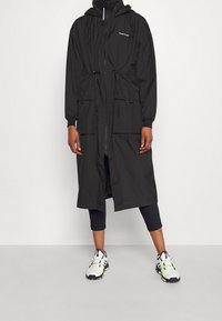 Abrigo de lluvia largo negro con capucha, cremallera y cordones. Cuenta con dos bolsillos frontales y puños elásticos. Se lleva sobre leggings oscuros y zapatillas.
