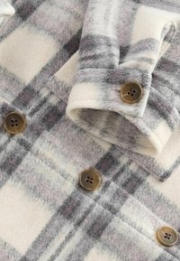 Koszula flanelowa w kratę w kolorze off-white z szarymi i lawendowymi wzorami. Posiada brązowe guziki oraz miękki, teksturowany materiał.