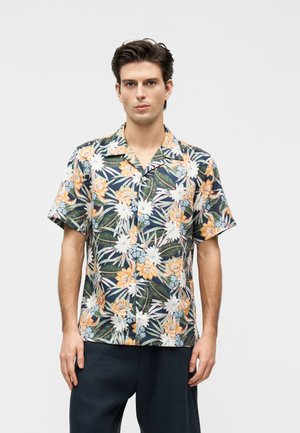 Jonge man draagt een donkerblauw shirt met korte mouwen met oranje, witte en groene bloemenpatronen, staand tegen een effen grijze achtergrond.