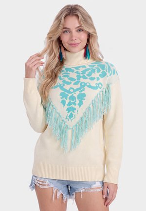 Lyshåret kvinde iført cremefarvet rullekravesweater med blåt blomsterprint og frynser, sat sammen med slidte denimshorts.