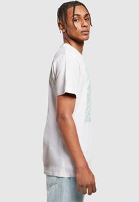 White cotton t-shirt featuring a light green palm leaf graphic print. Short sleeves, classic crew neck, and relaxed fit. Light blue jeans.