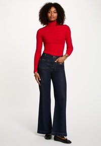 Crvena rebrasta turtleneck pulover uparen s tamnoplavim flared trapericama. Traperice imaju dekorativne džepove s metalnim detaljima. Crne ravne cipele.
