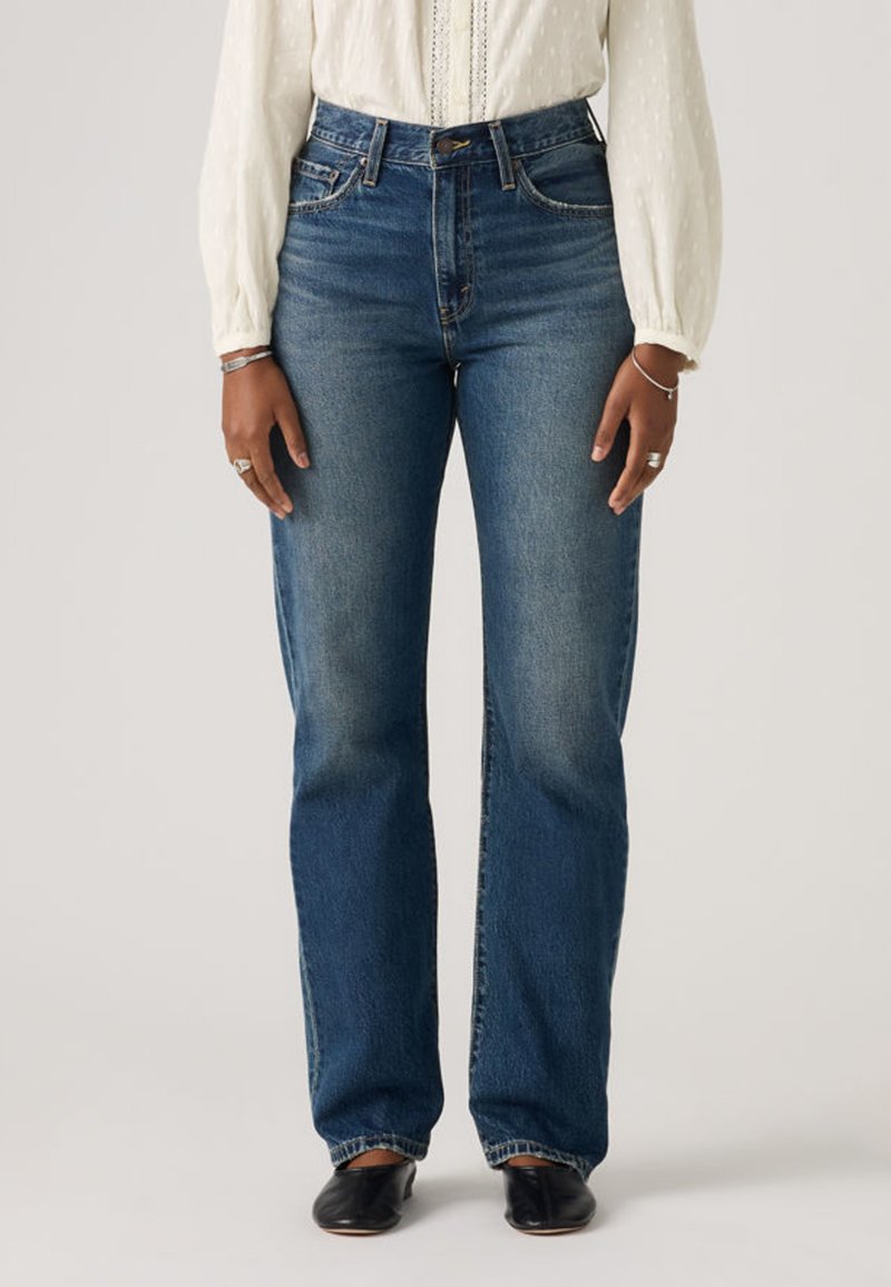 Γυναίκα που φοράει μπλε τζιν ψηλόμεσο με ίσια πόδια, λευκό μακρυμάνικο μπλουζάκι, μαύρα επίπεδα παπούτσια και ασημένια βραχιόλια και δαχτυλίδια.