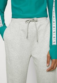 Calças de fato cinza-claro com cintura ajustável por cordão, bolsos laterais e textura suave. Combinadas com uma blusa de mangas longas em teal com padrões de folhas.