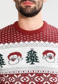 Czerwono-biała sweter dzianinowy z motywami świątecznymi: twarze św. Mikołaja, choinki i geometryczne wzory wzdłuż dolnego brzegu.