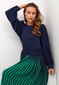 Bluză lungă de culoare albastru închis, cu umeri bombati, asortată cu o fustă plisată cu dungi verzi și albastre, având un elastic în talie.