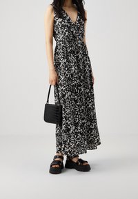 Robe longue à imprimé floral noir et blanc avec un décolleté en V, une taille cintrée et une jupe fluide. Associée à un sac à main noir texturé et des sandales épaisses.