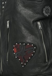 Jachetă din piele neagră cu fermoare argintii, un patch în formă de inimă cu accente roșii și cusături intricate, și studuri decorative în jurul patch-ului.