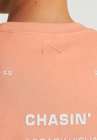 Persikinės spalvos džemperis, su baltu tekstu "CHASIN'" ir numeriais, matoma siuvimo detalė prie kaklo. Minkšta medvilnė.