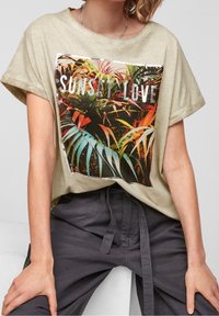 Luźna beżowa koszulka z żywym tropikalnym nadrukiem, na którym widnieje tekst "SUNSET LOVE" w srebrnym kolorze, zestawiona z ciemnoszarymi spodniami.