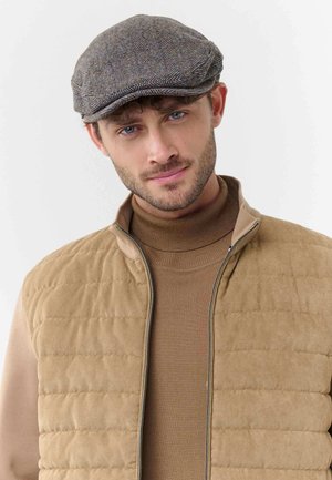 Young man with blue eyes wears a gray herringbone flat cap, tan quilted vest, and brown turtleneck sweater, facing forward with a slight smile.