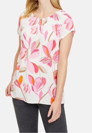 Vrouw draagt een witte korte mouwen blouse met roze, oranje en beige bloemenprint, gecombineerd met donkergrijze spijkerbroek, op een effen achtergrond.